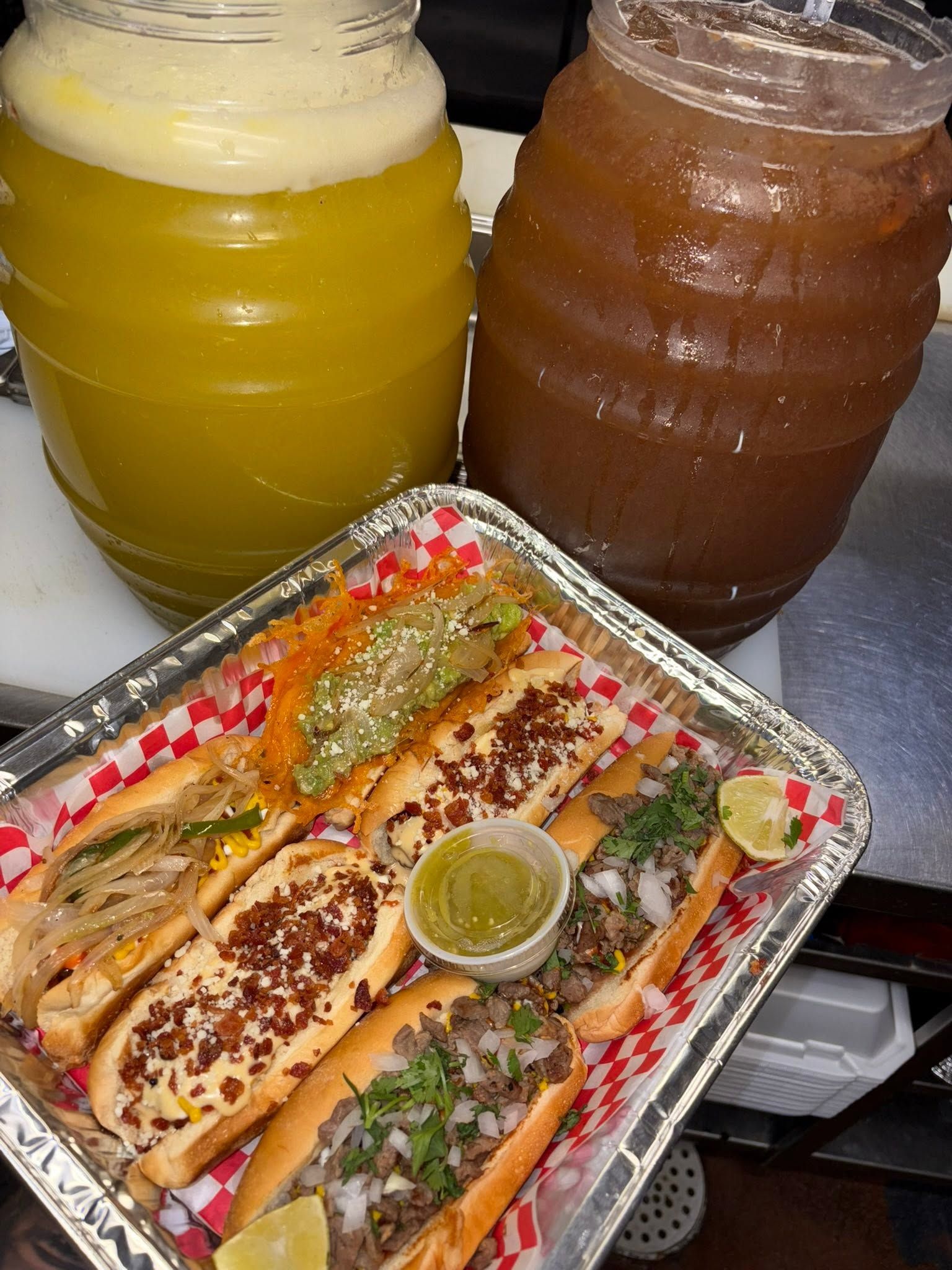 Two big jars of aguas frescas beside a tray of dogos