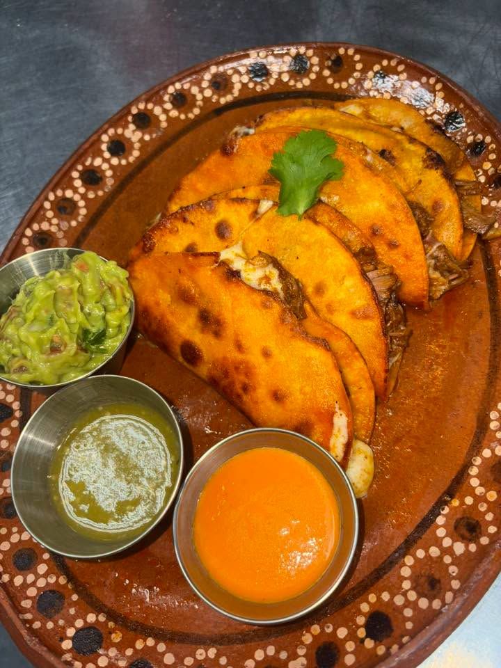 Four crisp quesa tacos on a barro plate with guacamole and cups of red and green salsa