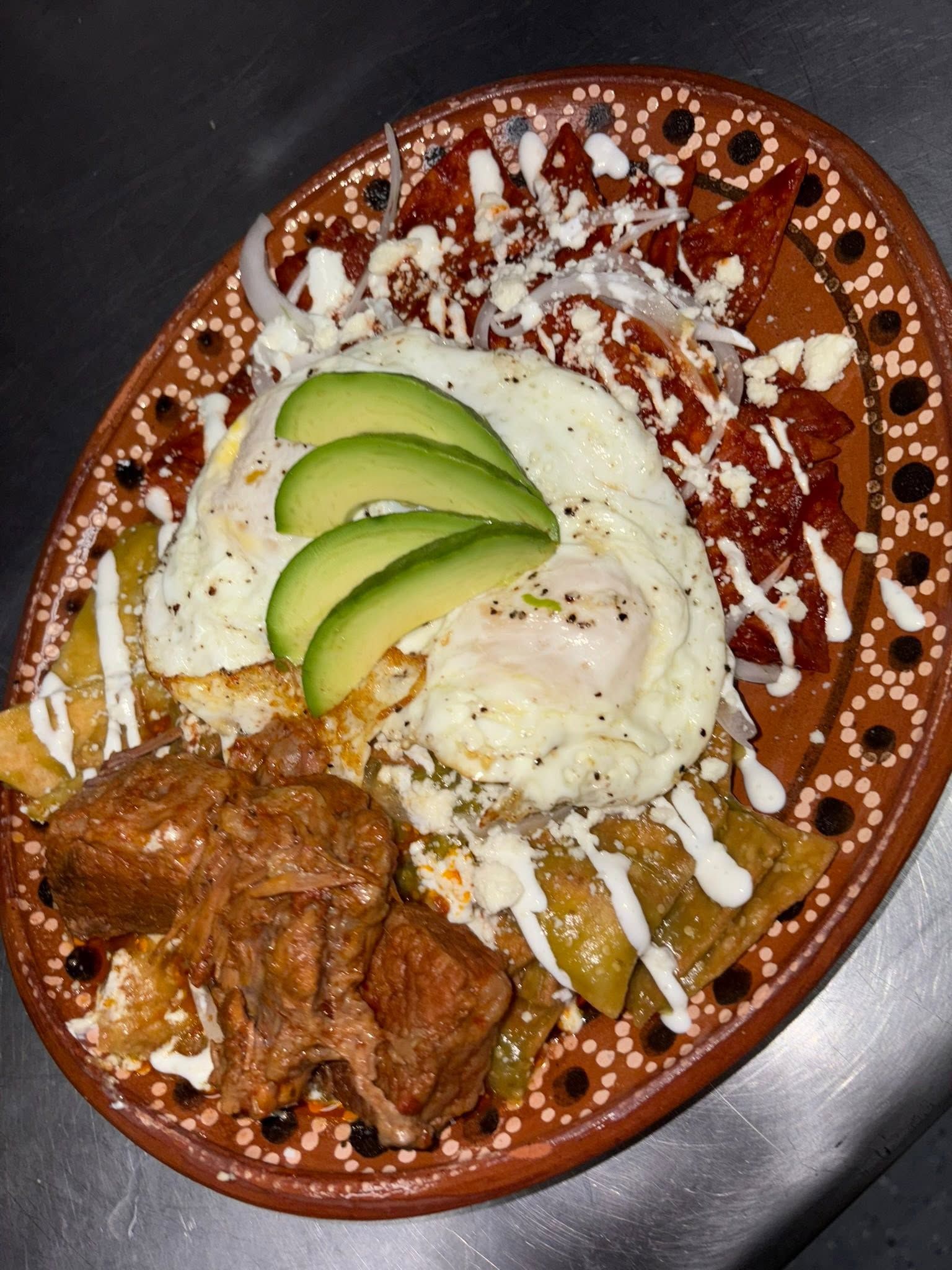 Chilaquiles under two fried eggs, sliced avocado and crema on a dotted barro plate