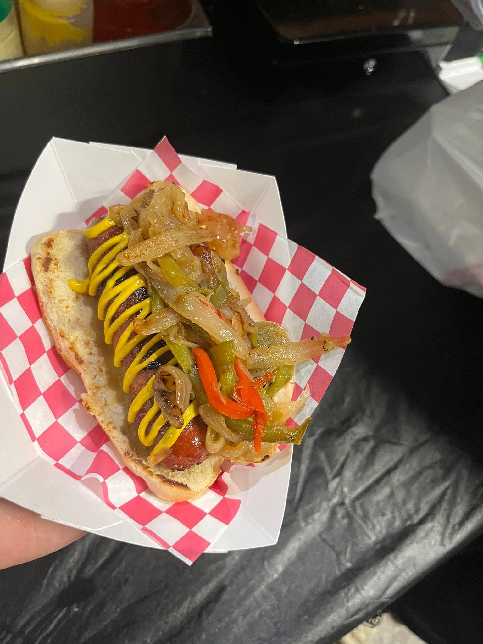 Grilled hot dog topped with mustard, grilled peppers, and onions in a paper basket