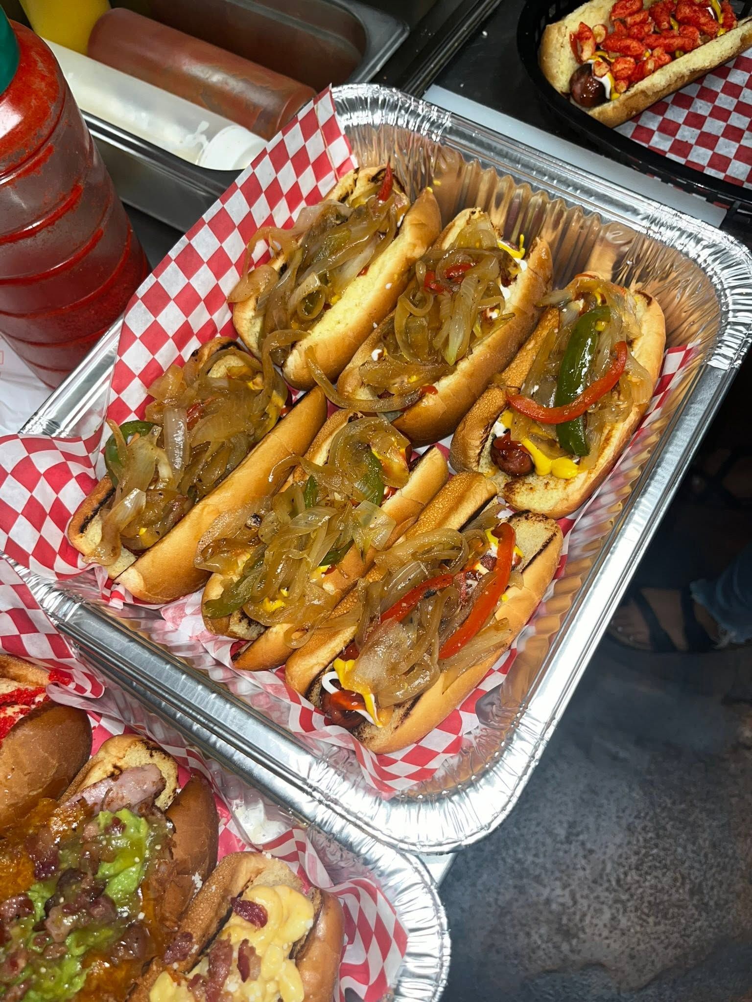 Foil tray of bacon-wrapped hot dogs topped with grilled onions and peppers