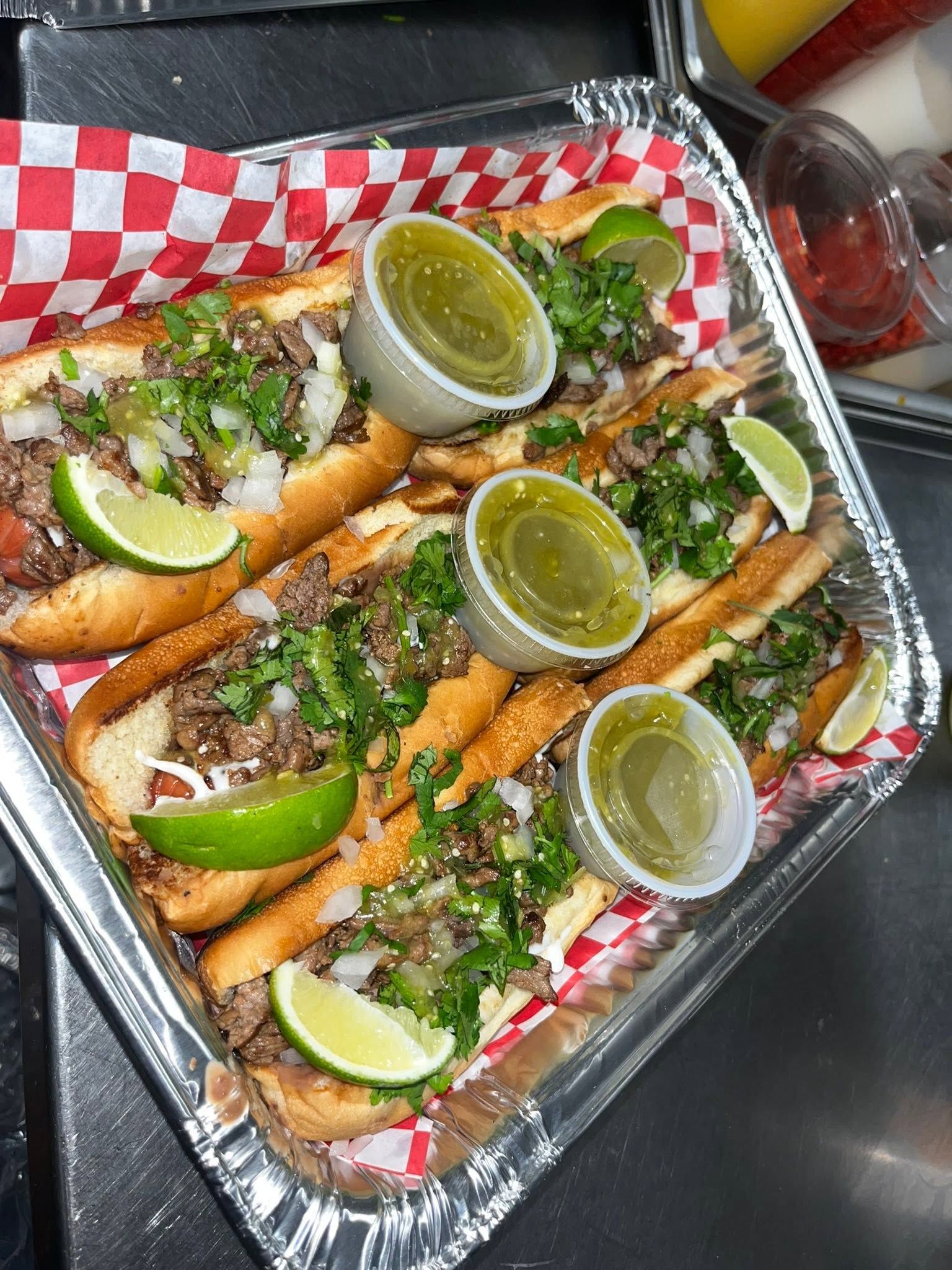A tray of asada dogos with cilantro and onion, lime wedges and salsa verde