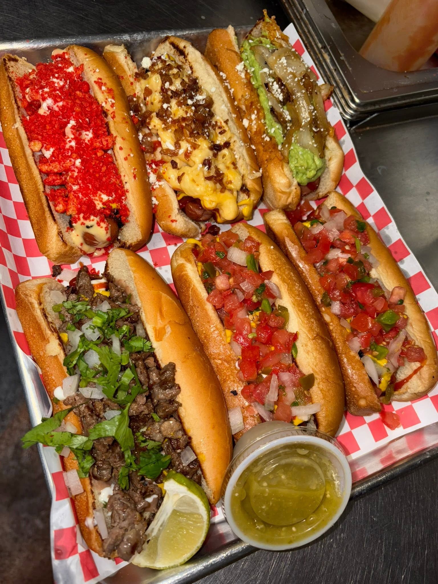 A foil tray of six loaded dogos: Hot Cheeto, mac and cheese with bacon, guacamole and pico