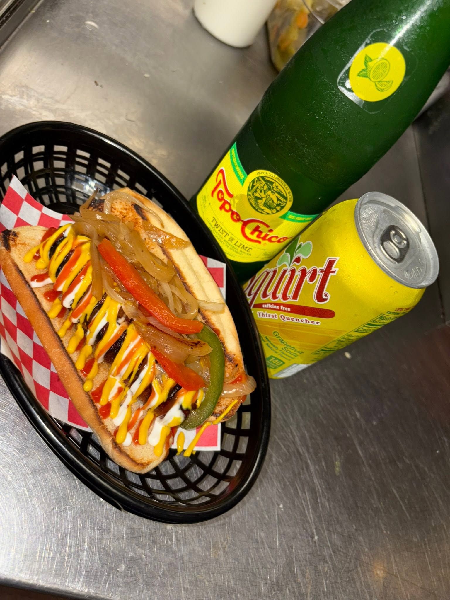 Bacon-wrapped dogo with grilled onions and peppers beside a Topo Chico bottle and Squirt can