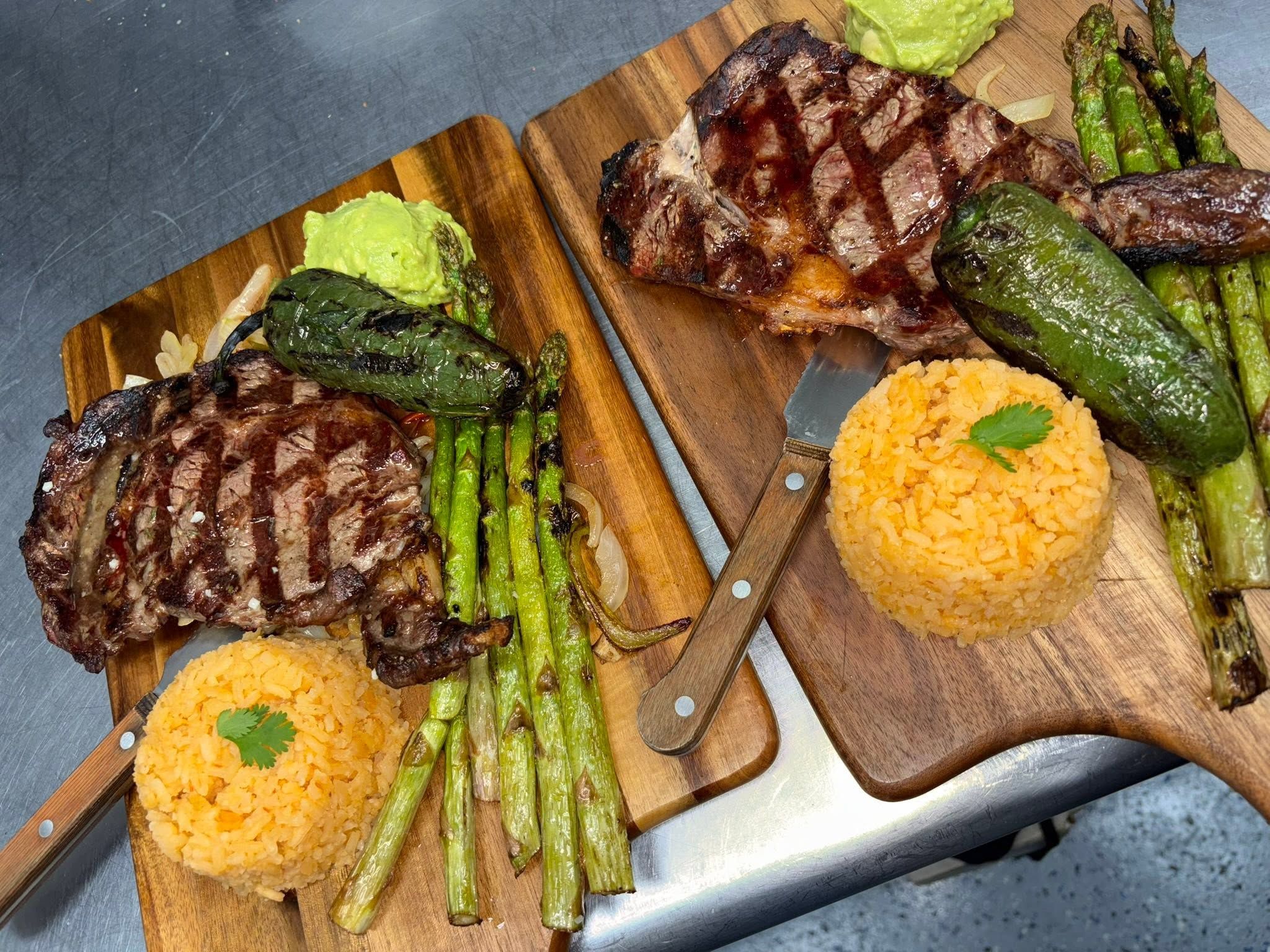 Two grilled ribeye steak boards with orange rice, charred asparagus and guacamole